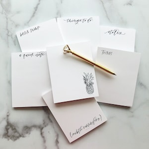 Puede incluir: Un conjunto de seis blocs de notas blancos con diferentes títulos, que incluyen "Brain Dump", "a quick note", "things to do", "notes", "THINGS" y "(subtle reminders)". También se puede ver una pluma dorada y una ilustración de piña.