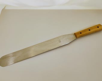 May include: A long, flat metal icing spatula with a rounded tip and a light-colored wooden handle secured by three rivets. The metal blade features a small stamped marking that reads US and 1945, indicating its origin and year of manufacture.