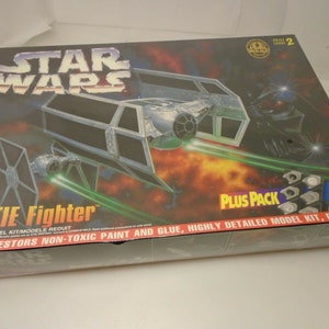 Puede incluir: Un kit de modelo de caza TIE de Star Wars con un paquete adicional de pintura y pegamento. La caja presenta un caza TIE en vuelo con un fondo rojo y naranja. La caja también incluye el texto "STAR WARS" y "TIE Fighter".