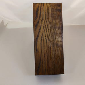 May include: A rectangular wooden block with a dark brown finish and a prominent, vertical wood grain pattern. The object has clean, straight edges and a smooth, flat surface, showcasing the natural texture and deep brown tones of the wood.