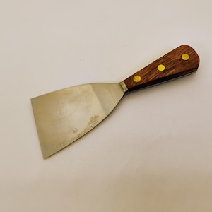 Puede incluir: Una espátula de metal con una hoja de acero ancha, plana y de color plateado, y un mango de madera. El mango está asegurado con tres remaches de color latón y muestra la palabra "Mabef" estampada en la madera cerca del refuerzo metálico.
