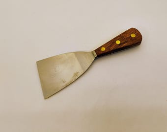 May include: A metal putty knife featuring a wide, flat, silver-colored steel blade and a wooden handle. The handle is secured with three brass-colored rivets and displays the word "Mabef" stamped into the wood near the metal bolster.