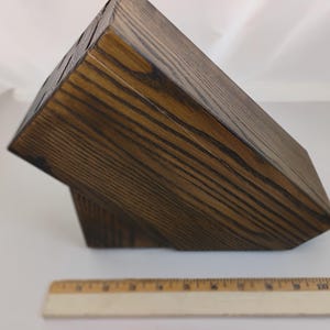 May include: A dark brown, stained wooden knife block with a visible wood grain pattern, featuring multiple slots on the top surface for knife storage. A wooden ruler is positioned in front, showing the block is approximately 11 inches long.