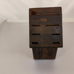 May include: A dark brown wooden knife block featuring a prominent wood grain pattern and multiple rectangular slots for storing kitchen knives. The front of the block displays a circular logo with the letters CASE embossed into the wood surface.