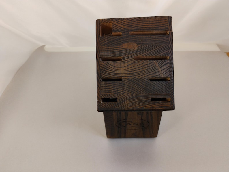 May include: A dark brown wooden knife block featuring a prominent wood grain pattern and multiple rectangular slots for storing kitchen knives. The front of the block displays a circular logo with the letters CASE embossed into the wood surface.