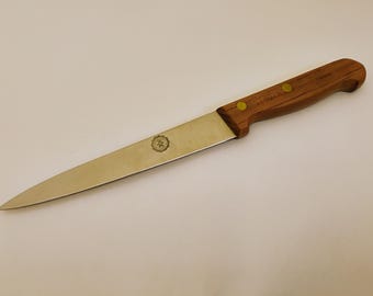 May include: A stainless steel knife with a wooden handle. The blade is long and slender, with a pointed tip. The handle is a warm brown color, secured with two brass rivets. A small emblem is visible on the blade.