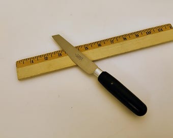 May include: A small knife with a silver blade and a black handle, resting on a wooden ruler. The ruler is marked with inch measurements. The knife appears to be a craft or utility tool.