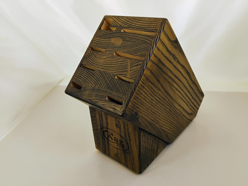 May include: A dark brown wooden knife block with a prominent wood grain pattern. The block features multiple angled slots for knife storage and displays the word CASE branded in black ink on the front face of the base section.