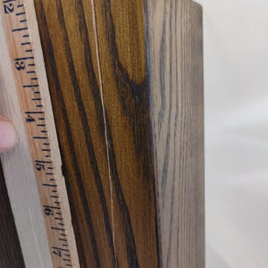 May include: A rectangular wooden block with a dark brown stained finish and visible wood grain, positioned next to a wooden ruler that shows a measurement of approximately 6.5 inches along the side of the object.