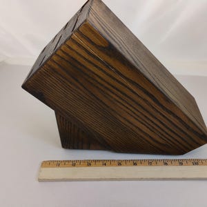 May include: A dark brown wooden knife block with a visible wood grain pattern and multiple slots for storage. A wooden ruler is positioned in front of the block, showing a length of approximately 11 inches for the base of the knife block.