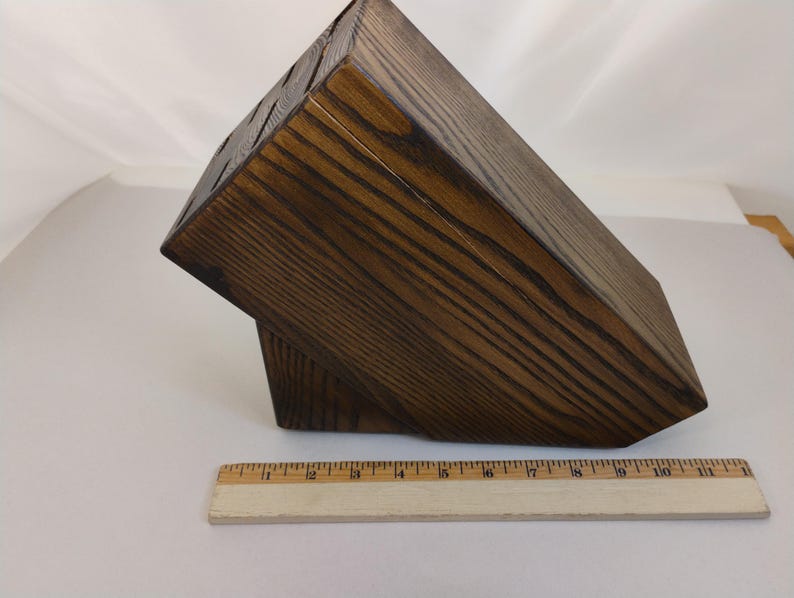 May include: A dark brown wooden knife block with a visible wood grain pattern and multiple slots for storage. A wooden ruler is positioned in front of the block, showing a length of approximately 11 inches for the base of the knife block.