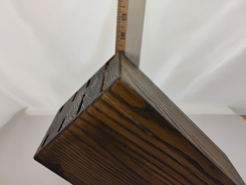 May include: A dark brown stained wooden knife block with visible wood grain and multiple rectangular slots on the top surface, positioned next to a metal ruler showing a measurement of approximately 8 inches in height for the block.