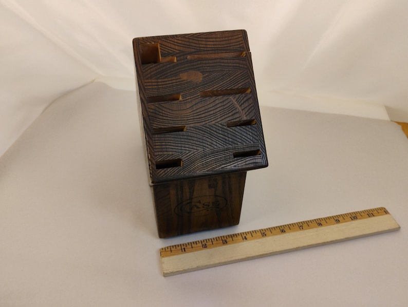 May include: A dark brown wooden knife block with a visible wood grain pattern and multiple slots for storage. A wooden ruler is positioned in front of the block, showing a length of approximately 11 inches for the ruler itself.
