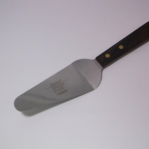 以下が含まれることがあります： 茶色の木製のハンドルが付いた銀色のケーキサーバー。サーバーには金色のリベットと、ブレードに「Basics Value Plus 71004152」という文字が印刷されています。