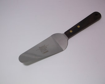 May include: A silver cake server with a brown wooden handle. The server has a gold-colored rivet and the text "Basics Value Plus 71004152" printed on the blade.