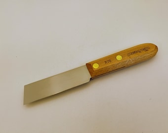 May include: A putty knife with a flat, rectangular metal blade and a light brown wooden handle. The handle is secured with two brass-colored rivets and features the text DEXTER U.S.A. and the model number X70 clearly printed on the wood surface.