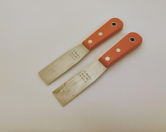 May include: Two putty knives with orange handles and silver metal blades. Each blade is stamped with the text R. Murphy, Ayer, MA, 5-1 1/4-S. The handles are secured with three metal rivets and feature a circular hole at the end for hanging.