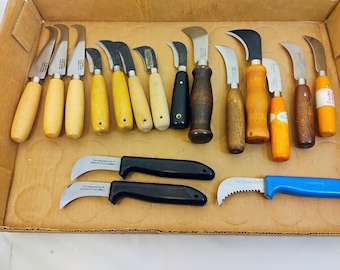 May include: A collection of fifteen curved knives with various handle materials including light wood, dark wood, black plastic, and blue plastic. Several blades feature the text The Organic Co. and Forged in Columbia. The knives are arranged in a cardboard box.