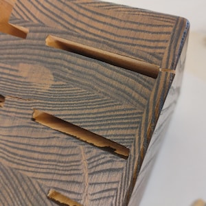 May include: A wooden knife block featuring a dark brown and tan wood grain pattern with multiple horizontal slots for storing kitchen knives. The block has a rectangular shape with visible wood texture and natural grain lines throughout the surface.