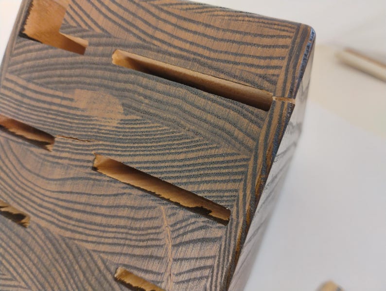May include: A wooden knife block featuring a dark brown and tan wood grain pattern with multiple horizontal slots for storing kitchen knives. The block has a rectangular shape with visible wood texture and natural grain lines throughout the surface.