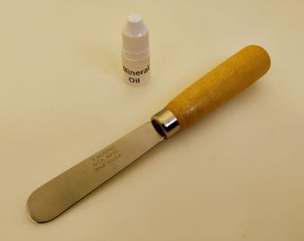 Könnte beinhalten: Ein Edelstahlspatel mit Holzgriff und eine kleine Flasche Mineralöl. Der Spatel ist mit "R. MURPHY AYER, MASS MADE IN U.S.A." gekennzeichnet. Die Flasche ist weiß mit weißem Deckel und dem Text "Mineral Oil".