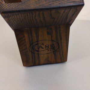 May include: A dark brown wooden knife block with a tapered, trapezoidal shape and a visible wood grain texture. The front face features the black printed text CASE XX inside an oval border, with a registered trademark symbol to the right.
