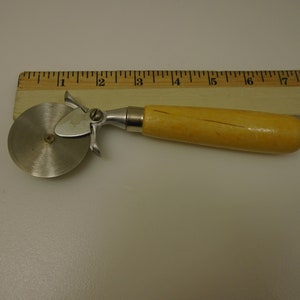 Puede incluir: Un cortador de pizza plateado con mango de madera. El cortador tiene una cuchilla redonda con una pequeña cuchilla puntiaguda en el lateral. El mango mide aproximadamente 15 cm de largo.