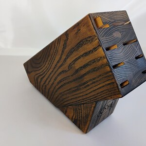 May include: A dark brown wooden knife block with a prominent wood grain pattern. The block features an angled top surface with six rectangular slots for storing kitchen knives and a small circular logo stamped on the lower side panel.