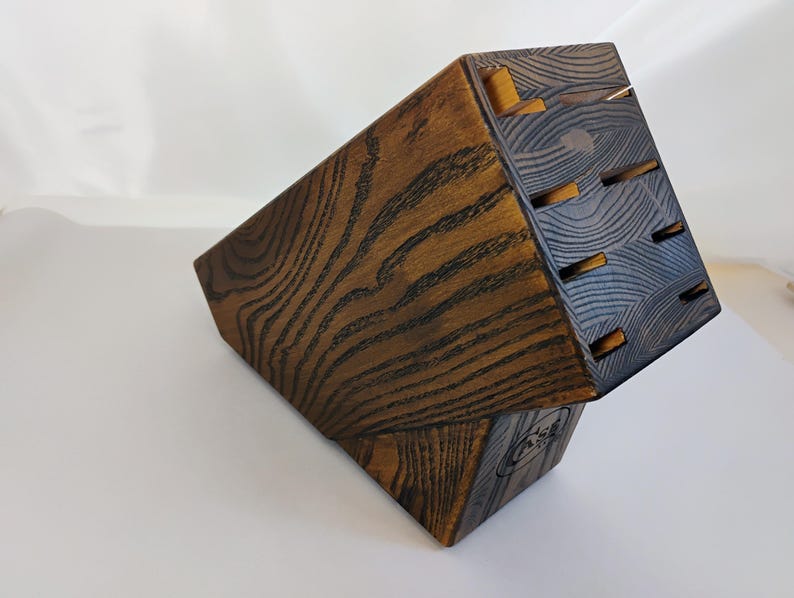 May include: A dark brown wooden knife block with a prominent wood grain pattern. The block features an angled top surface with six rectangular slots for storing kitchen knives and a small circular logo stamped on the lower side panel.