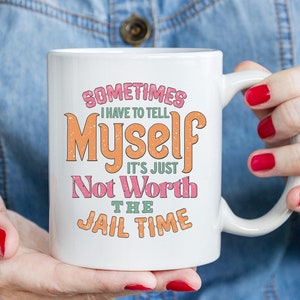 May include: White ceramic mug with the text "Sometimes I have to tell Myself it's just Not Worth the Jail Time" in retro-style lettering. The mug is held by a person with red nail polish.