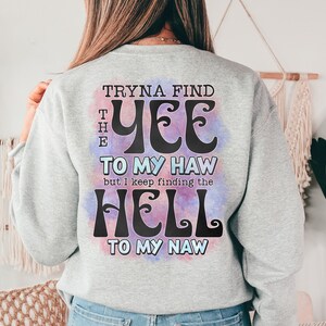 May include: A light gray sweatshirt with the text "Tryna Find The Yee To My Haw but I keep finding the Hell To My Naw" in a whimsical font. The text is over a watercolor-style background with purple and blue hues. The sweatshirt is a casual style.