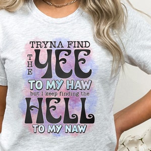May include: Light gray t-shirt with the text "Tryna Find The Yee To My Haw but I Keep Finding The Hell To My Naw" in black, with a watercolor effect in pink and blue. The shirt is a casual wear item.
