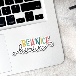 May include: A colorful sticker with the text "BE A NICE human" in a whimsical font. The sticker is on a white laptop.