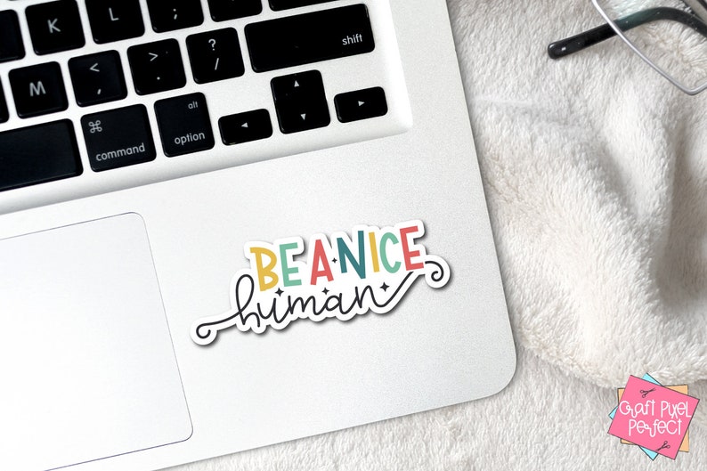 May include: A colorful sticker with the text "BE A NICE human" in a whimsical font. The sticker is on a white laptop.