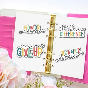 May include: Four colorful motivational stickers with positive messages: "Now or never", "Never give up", "Make a difference", and "Be a nice human". The stickers are placed on a pink binder with a gold ring mechanism.