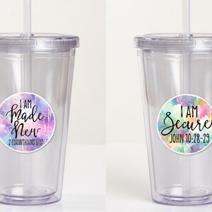 May include: Two clear plastic tumblers with lids and straws. The tumblers have colorful watercolor-style stickers with inspirational quotes and bible verses. The left tumbler has a sticker that says "I AM Made New 2 Corinthians 5:17". The right tumbler has a sticker that says "I AM Secure John 10:28-29".