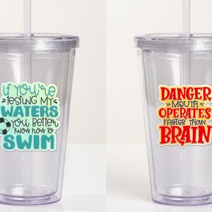 May include: Two clear plastic tumblers with reusable straws. The tumbler on the left has a teal and blue graphic that says "If you're testing my waters you better know how to swim". The tumbler on the right has a red graphic that says "Danger >>Mouth Operates Faster Than Brain".