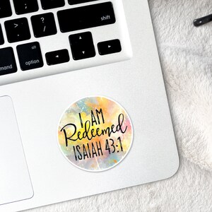 May include: A round sticker with a watercolor background in shades of yellow, orange, pink, and blue. The sticker features the text "I AM Redeemed ISAIAH 43:1" in black.