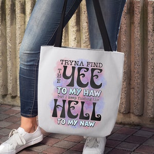 May include: White tote bag with black handles featuring the text "Tryna find the Yee to my Haw but I keep finding the Hell to my Naw" in a whimsical font. The design includes pink and blue watercolor-style accents.