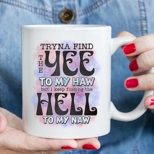 May include: White ceramic mug with the text "Tryna find the yee to my haw but I keep finding the hell to my naw" in black lettering. The text is set against a watercolor-style background with pink and purple hues.