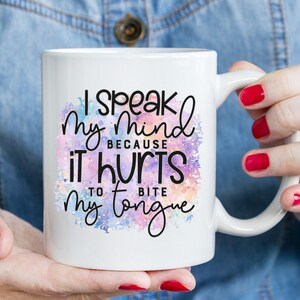 K&ouml;nnte beinhalten: Eine wei&szlig;e Keramiktasse mit einem Aquarellhintergrund in Rosa-, Lila- und Blaut&ouml;nen. Die Tasse tr&auml;gt einen schwarzen Text mit der Aufschrift "Ich sage meine Meinung, weil es weh tut, sich auf die Zunge zu bei&szlig;en."