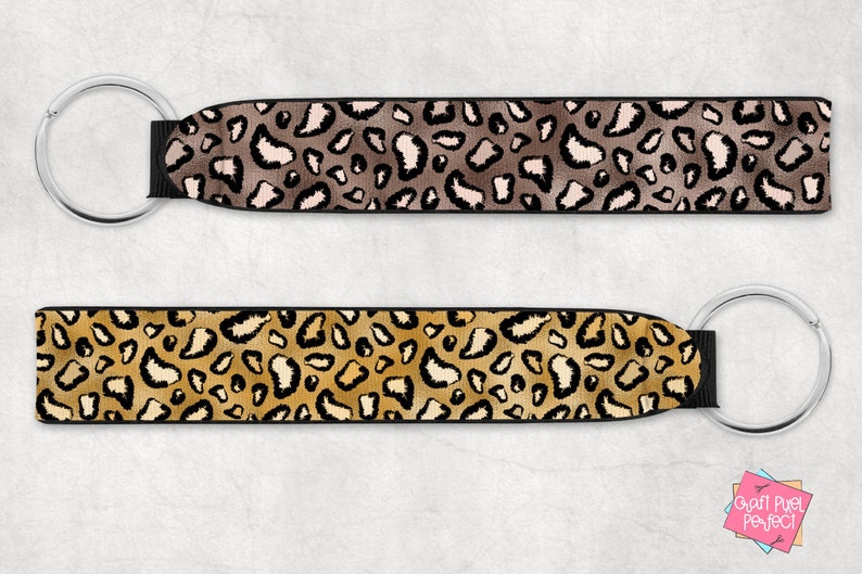 May include: Two keychains with black fabric and silver key rings. The fabric has a leopard print pattern. One keychain has a brown and beige leopard print, and the other has a gold and beige leopard print.