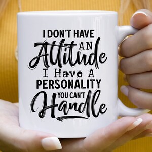 Puede incluir: Taza de cer&aacute;mica blanca con un dise&ntilde;o de texto negro. La taza presenta la frase "I DON'T HAVE AN Attitude I Have A PERSONALITY YOU CAN'T Handle" en una fuente audaz y estilizada. La taza est&aacute; sostenida por una persona.