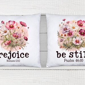 May include: Two white pillow covers with floral designs and inspirational quotes. The left pillow features pink and white flowers with the quote "rejoice" and the scripture reference "Romans 12:12". The right pillow features the same floral design with the quote "be still" and the scripture reference "Psalm 46:10".