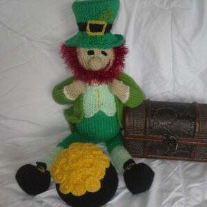 K&ouml;nnte beinhalten: Eine gr&uuml;ne und rote gestrickte Koboldpuppe, die einen gr&uuml;nen Hut mit einer goldenen Schnalle tr&auml;gt. Die Puppe h&auml;lt einen Topf mit Gold.