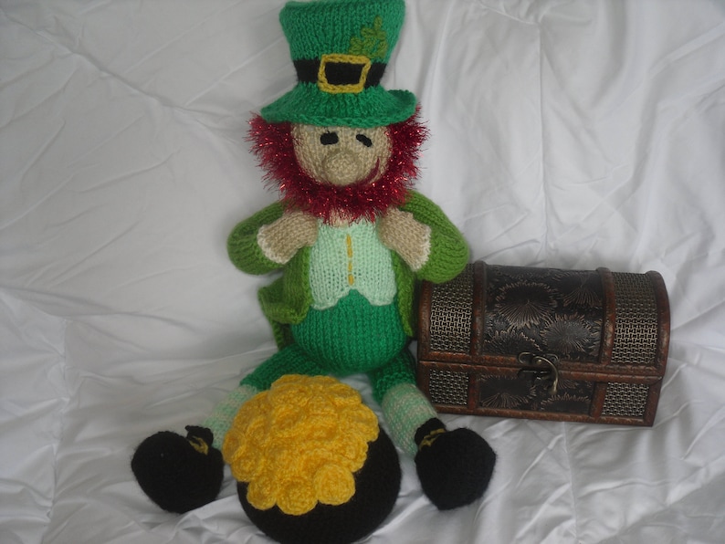K&ouml;nnte beinhalten: Eine gr&uuml;ne und rote gestrickte Koboldpuppe, die einen gr&uuml;nen Hut mit einer goldenen Schnalle tr&auml;gt. Die Puppe h&auml;lt einen Topf mit Gold.