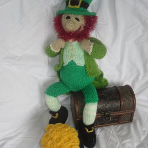 K&ouml;nnte beinhalten: Eine gr&uuml;ne gestrickte Koboldpuppe, die einen gr&uuml;nen Hut mit einer goldenen Schnalle und einen roten Bart tr&auml;gt. Die Puppe sitzt auf einer braunen Holzkiste mit einem goldenen Schloss. Die Puppe h&auml;lt einen Topf mit Goldm&uuml;nzen.