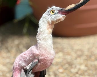 May include: A handcrafted bird figurine featuring a long, slender neck with soft white and pale pink fur. The bird has a prominent, elongated beak with a black tip and a textured base, along with a single visible yellow eye with a dark pupil.