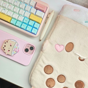 May include: A pink phone case with a cartoon boba tea character on the front. The case is lying on a white surface next to a keyboard and a plush brown and white pouch with a heart and boba tea design.
