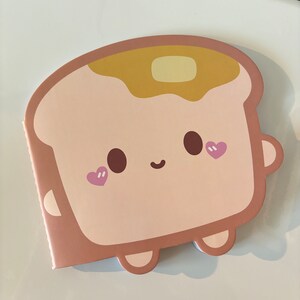 May include: A notepad shaped like a slice of toast with a smiling face, heart-shaped blush, and a pat of butter on top. The notepad has a light brown color with a darker brown outline and details.